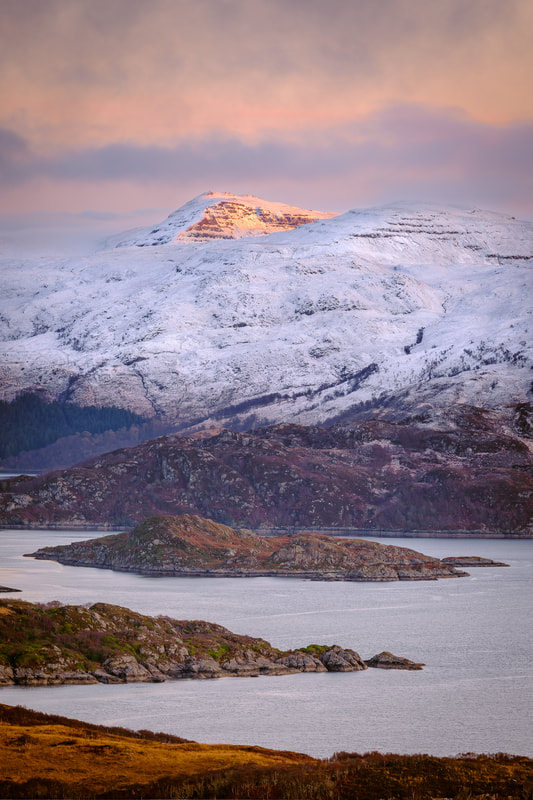 The snow-capped peak of Beinn Iadain lit up with golden light of a sunset with Beinn Ghormaig, the Isle Carna and the Isle of Risga in the foreground | Morvern Scotland