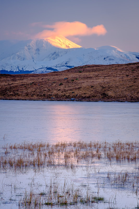 First light of a winter morning hitting a snow-covered Ben Nevis with its peak capped with cloud viewed from Lochan Doire a' Bhraghaid | Ardgour Scotland | Steven Marshall Photography