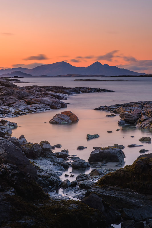 Photographic print of the rugged peaks of the Rùm Cuillin silhouetted against the orange of a dusk sky, viewed immediately after sunset from amongst the rocky shoreline of Inbhir Allt na Luachair. Image of Ardnamurchan on the West Highland Peninsulas, Scotland.