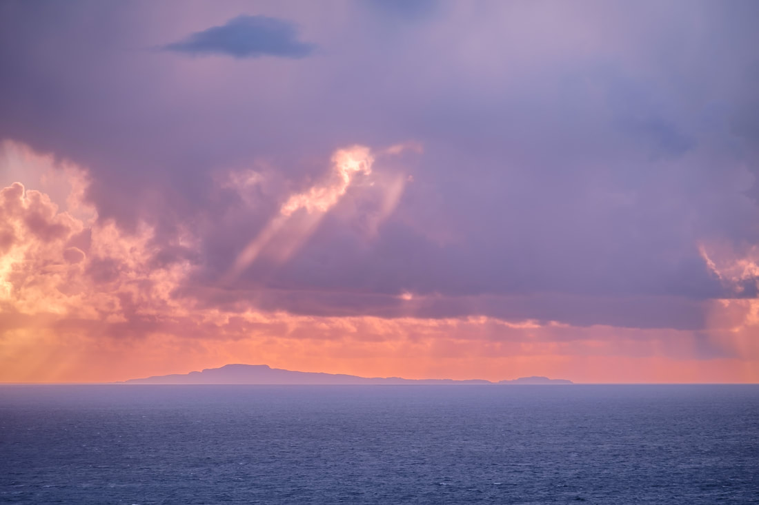 The Isle of Muck set against a pink sky with crepuscular rays breaking through the cloud above it | Moidart Scotland
