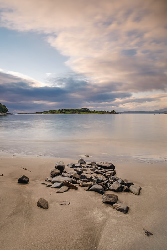 Samalaman Beach at high tide with a pile of rocks in the sand just above the water line with clouds parting to reveal blue sky | Moidart Scotland