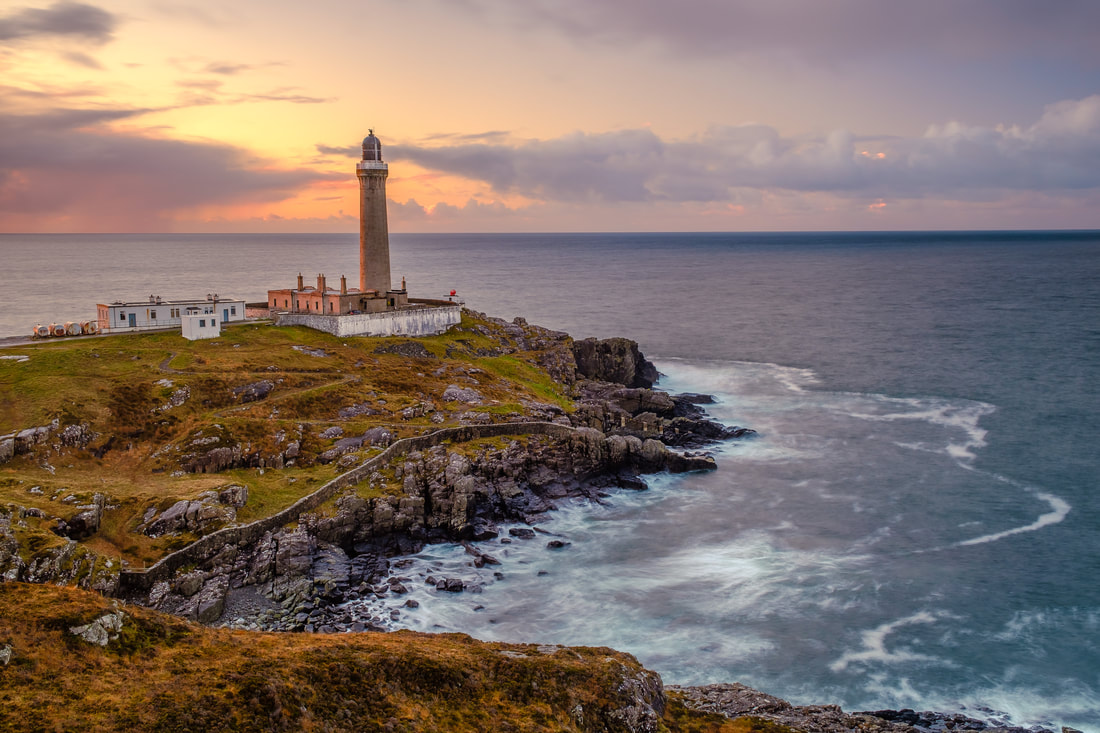 Ardnamurchan Lighthouse on a blustery afternoon with light from the setting sun falling on its side and waves crashing on the rocks beneath it| Ardnamurchan Scotland