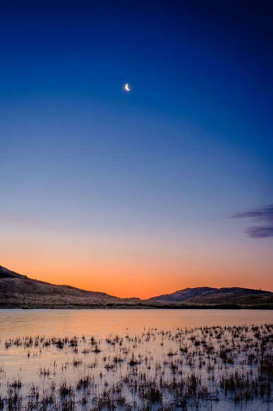 A waning crescent moon above Lochan Doire a' Bhraghaid and the orange of a rising sun colouring its frozen surface and the sky above the hills beyond | Ardgour Scotland