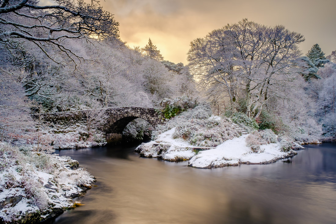 The old bridge over the River Shiel at dawn after a fresh fall of overnight snow, with the golden light of the rising sun colouring the sky and the river | Moidart Scotland