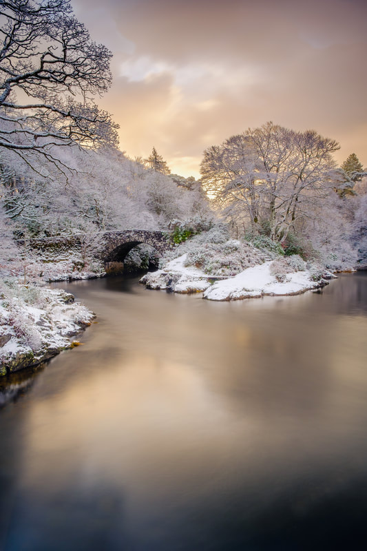 The old bridge over the River Shiel at dawn after a fresh fall of overnight snow, with the golden light of the rising sun colouring the sky and the river | Moidart Scotland