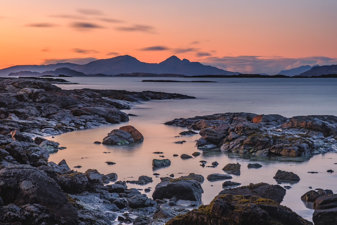 Photographic print of the rugged peaks of the Rùm Cuillin silhouetted against the orange of a dusk sky, viewed immediately after sunset from amongst the rocky shoreline of Inbhir Allt na Luachair. Image of Ardnamurchan on the West Highland Peninsulas, Scotland.