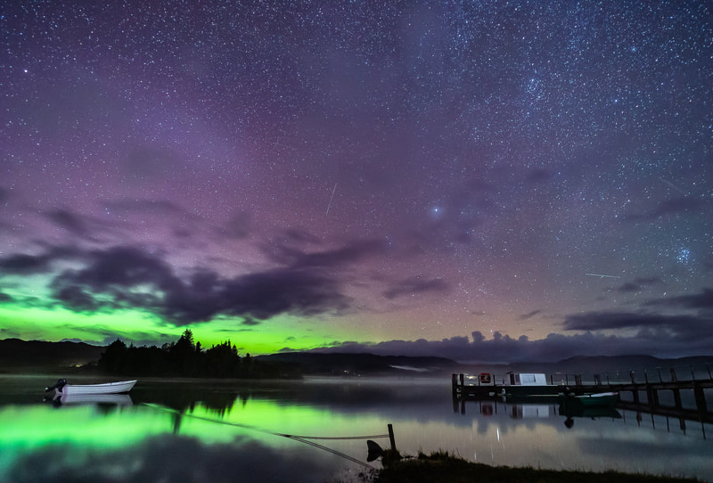 A small day boat moored up in Loch Shiel at the jetty in Acharacle floats on water that has been lit green by the aurora in the sky beyond the hills to the north.